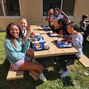 Five students eating lunch at a picnic table and holding up their apples.